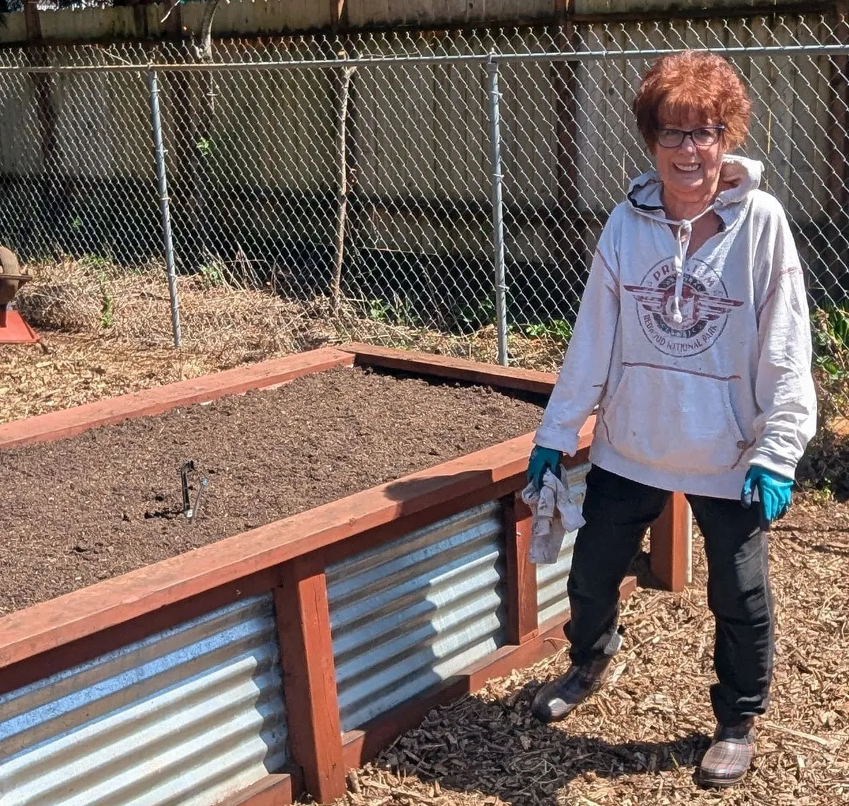 A smiling person in gardening gloves beside a raised garden bed.
